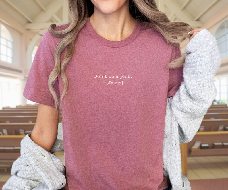 Peut inclure: Un t-shirt rose poudr&eacute; avec l'inscription blanche "Don't be a jerk. -[Jesus]" sur le devant. Le t-shirt est de style col rond et semble &ecirc;tre fait d'une mati&egrave;re douce. L'arri&egrave;re-plan montre un int&eacute;rieur d'&eacute;glise.