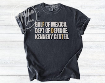 Könnte beinhalten: Ein anthrazitfarbenes Kurzarm-T-Shirt mit weißem Text, der GULF OF MEXICO. DEPT OF DEFENSE. KENNEDY CENTER. lautet. Die Buchstaben F, D und T sind in einem matten Goldton hervorgehoben und bilden eine vertikale Ausrichtung in der Mitte.