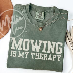 May include: Olive green t-shirt with the text "MOWING IS MY THERAPY" in white block letters. A gold necklace with a small pendant rests on the shirt. A brown hat and dried plant are in the background.
