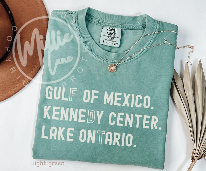 May include: A light green short-sleeve t-shirt with a crew neckline. The shirt features white text that reads GULF OF MEXICO. KENNEDY CENTER. LAKE ONTARIO. with the letters F, D, and T highlighted in a different font style on the front.