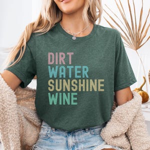 May include: Dark green t-shirt with the words "DIRT WATER SUNSHINE WINE" in pastel colours. The shirt is made of a soft material and is a casual style.