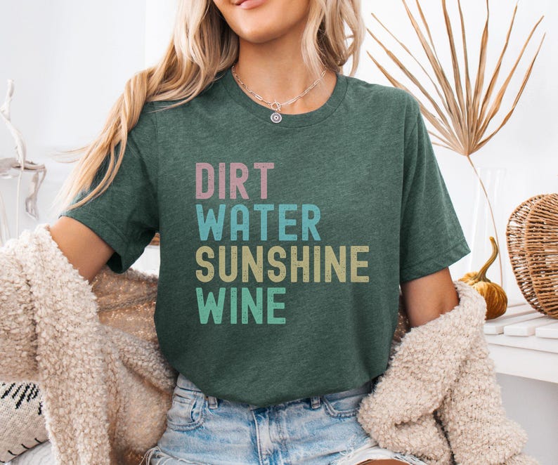 May include: Dark green t-shirt with the words "DIRT WATER SUNSHINE WINE" in pastel colours. The shirt is made of a soft material and is a casual style.