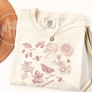 May include: Cream-colored t-shirt with a pink illustration of tea-related elements, including berries, a teacup, a butterfly, and the words "tea time." A gold necklace and a brown hat are also visible.