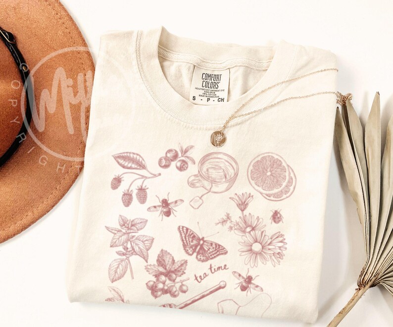 May include: Cream-colored t-shirt with a pink illustration of tea-related elements, including berries, a teacup, a butterfly, and the words "tea time." A gold necklace and a brown hat are also visible.