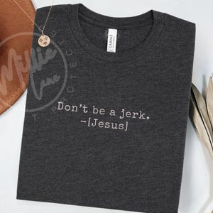 Peut inclure: T-shirt gris fonc&eacute; avec l'inscription blanche "Don't be a jerk. -[Jesus]" brod&eacute;e. Le t-shirt a un col rond et une &eacute;tiquette "CANVAS". Un collier dor&eacute; et un chapeau marron sont en arri&egrave;re-plan.