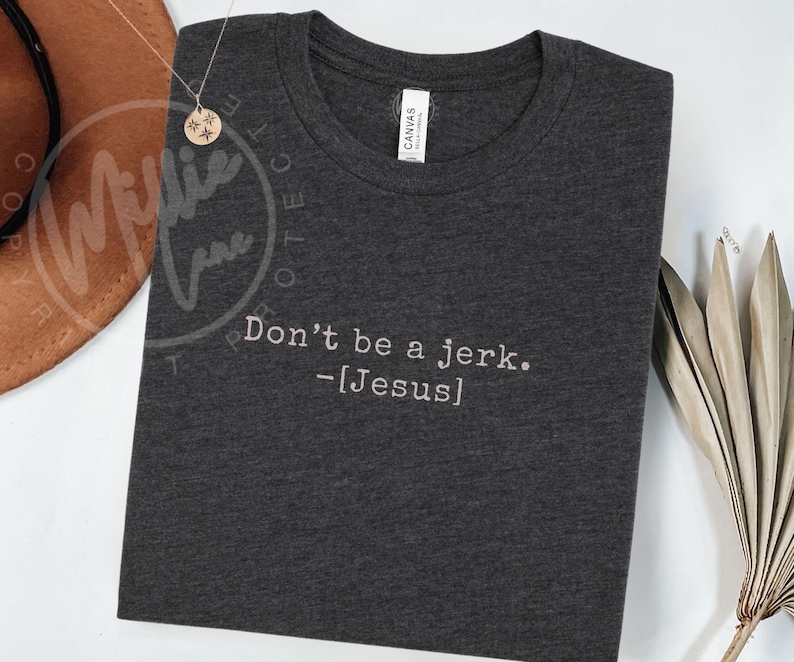 Peut inclure: T-shirt gris fonc&eacute; avec l'inscription blanche "Don't be a jerk. -[Jesus]" brod&eacute;e. Le t-shirt a un col rond et une &eacute;tiquette "CANVAS". Un collier dor&eacute; et un chapeau marron sont en arri&egrave;re-plan.