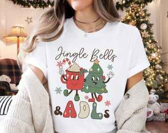 May include: White t-shirt with a Christmas-themed graphic. The graphic features a red mug with a candy cane and a green Christmas tree, both with smiling faces. The text "Jingle Bells & ADLs" is printed below the graphic.