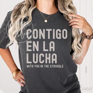 May include: Dark gray t-shirt with the text "CONTIGO EN LA LUCHA" in large, light gray letters. Below, the English translation reads "WITH YOU IN THE STRUGGLE". The shirt has short sleeves and a crew neck.
