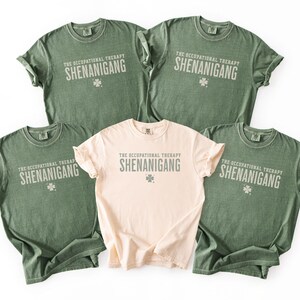 May include: Five t-shirts, four in olive green and one in a light peach color. Each shirt has the text "THE OCCUPATIONAL THERAPY SHENANIGANG" printed in a light grey color, with a small four-leaf clover graphic.