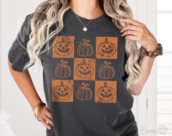 Puede incluir: Camiseta gris oscuro con una cuadrícula de nueve cuadrados naranjas, cada uno con un diseño diferente de calabaza de Halloween. Las calabazas tienen varias caras talladas. La camiseta es de estilo casual y cómodo, perfecta para Halloween.