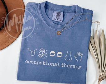 May include: A blue Comfort Colors t-shirt with white graphic icons of the tongue, ear, eyes, nose, and hand, with the words "occupational therapy" below. A silver necklace with a small pendant is on the shirt.