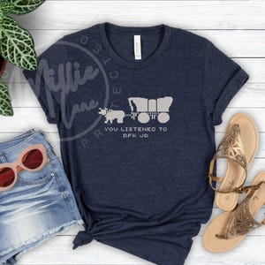 Puede incluir: Camiseta azul marino con un gr&aacute;fico de una carreta cubierta tirada por un oso, con el texto "YOU LISTENED TO RFK JR" en blanco. La camiseta se combina con pantalones cortos de mezclilla y gafas de sol.