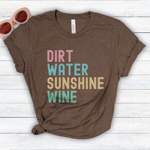 May include: A brown t-shirt featuring the words "DIRT WATER SUNSHINE WINE" in pastel shades. The tee is folded on a white wooden surface, with pink sunglasses in the top left corner.