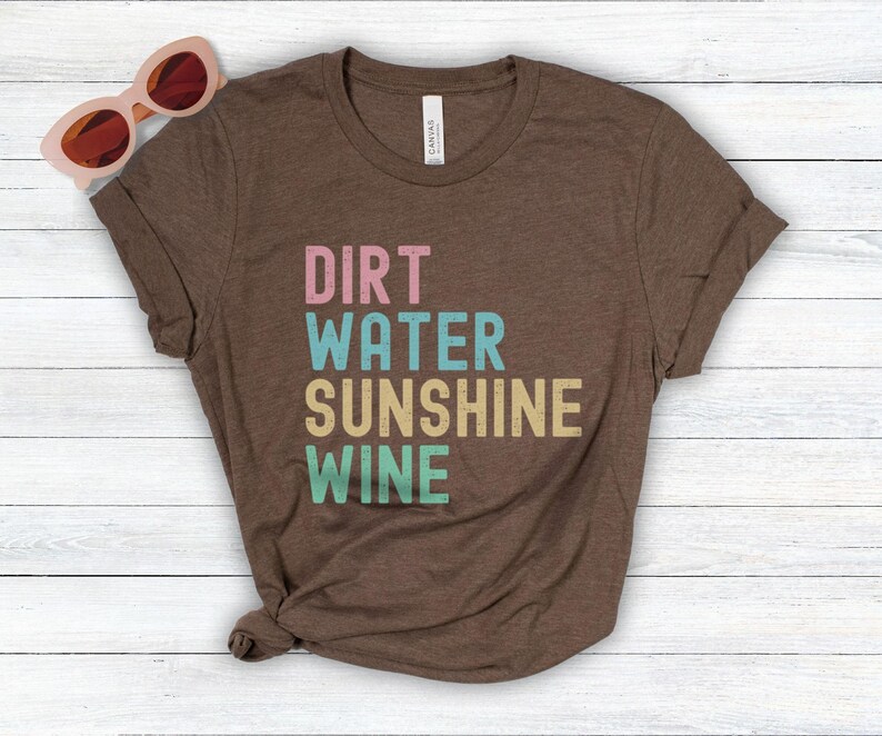 May include: A brown t-shirt featuring the words "DIRT WATER SUNSHINE WINE" in pastel shades. The tee is folded on a white wooden surface, with pink sunglasses in the top left corner.
