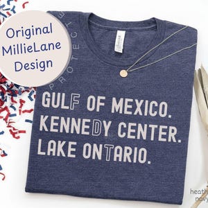May include: A heather navy t-shirt featuring white text that reads GULF OF MEXICO, KENNEDY CENTER, and LAKE ONTARIO. The shirt has a crew neckline and a small white brand tag at the inner collar. A thin silver chain necklace rests on the fabric.