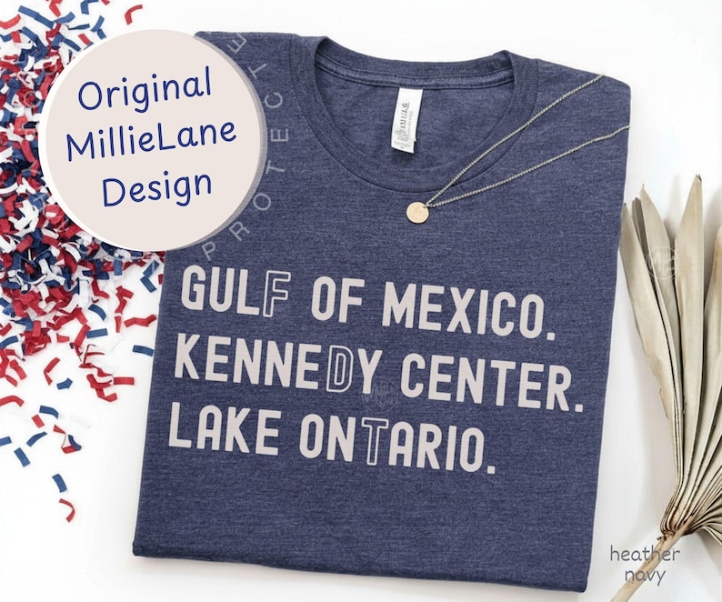 May include: A heather navy t-shirt featuring white text that reads GULF OF MEXICO, KENNEDY CENTER, and LAKE ONTARIO. The shirt has a crew neckline and a small white brand tag at the inner collar. A thin silver chain necklace rests on the fabric.