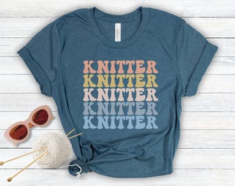 May include: A teal blue t-shirt with the word "Knitter" repeated five times in a rainbow gradient pattern. The text is in a retro font style.