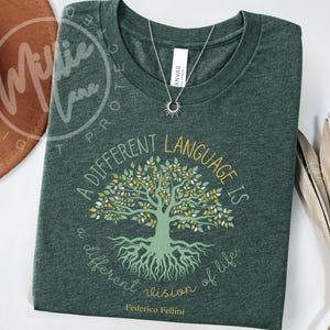 May include: Green t-shirt with a round neckline, featuring a tree graphic and the quote "A different language is a different vision of life" by Federico Fellini. A silver necklace with a sun pendant is visible.