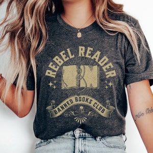 K&ouml;nnte beinhalten: Ein dunkelgraues T-Shirt mit einem Vintage-Grafikdesign. Die Grafik zeigt ein Buch mit einem Band-Lesezeichen, umgeben von Sternen und Lichtstrahlen. Der Text "Rebel Reader Banned Books Club" ist in einer dekorativen Schrift geschrieben.