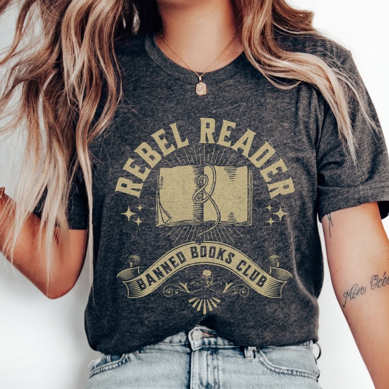 K&ouml;nnte beinhalten: Ein dunkelgraues T-Shirt mit einem Vintage-Grafikdesign. Die Grafik zeigt ein Buch mit einem Band-Lesezeichen, umgeben von Sternen und Lichtstrahlen. Der Text "Rebel Reader Banned Books Club" ist in einer dekorativen Schrift geschrieben.