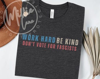 May include: Dark gray t-shirt with the text "WORK HARD BE KIND DON'T VOTE FOR FASCISTS" in blue, beige, and red. The shirt is made of a soft material and has a crew neck. A gold necklace is visible in the upper left corner.