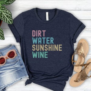 May include: A navy blue t-shirt displaying the words "DIRT WATER SUNSHINE WINE" in a stacked, multi-coloured font. The image includes denim shorts, sunglasses, and sandals, creating a relaxed, summery aesthetic.