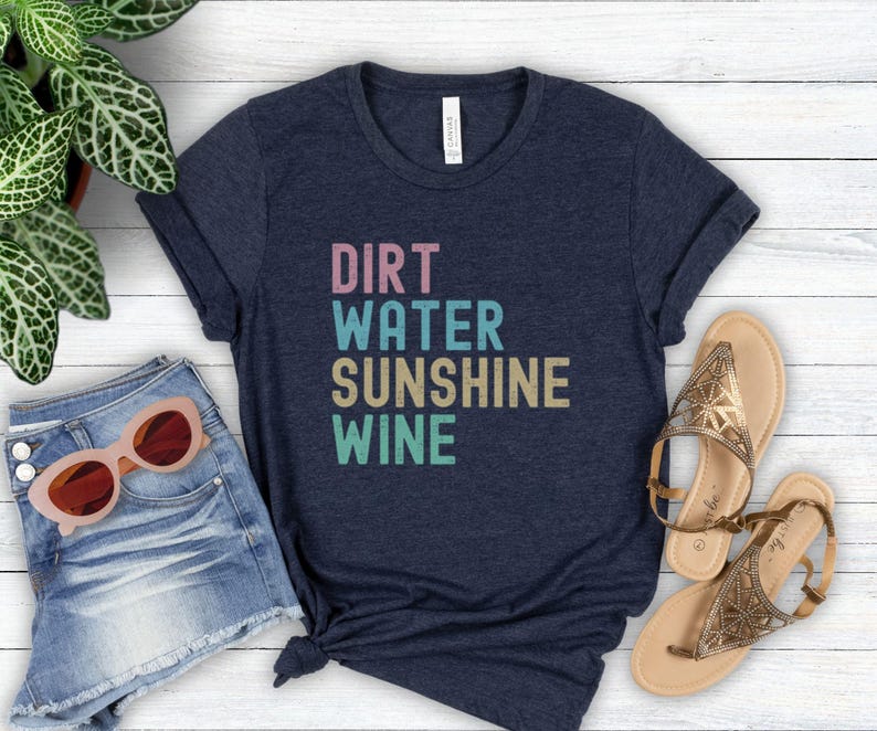 May include: A navy blue t-shirt displaying the words "DIRT WATER SUNSHINE WINE" in a stacked, multi-coloured font. The image includes denim shorts, sunglasses, and sandals, creating a relaxed, summery aesthetic.