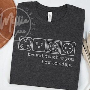 May include: Dark gray t-shirt with a white graphic of international electrical outlet designs. The text "travel teaches you how to adapt" is printed below the outlets. The shirt is made of a soft material.
