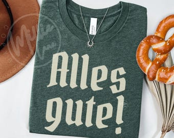 Op de afbeelding: Een donkergroen t-shirt met de woorden "Alles Gute!" in een crèmekleurige, vette letter. Een zilveren ketting met een zonnehanger is zichtbaar. Een pretzel en een hoed staan op de achtergrond.