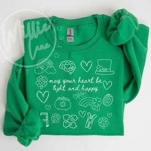 May include: A green sweatshirt with white St. Patrick's Day themed doodles, including a rainbow, hearts, and a leprechaun hat. The text "may your heart be light and happy" is also featured.