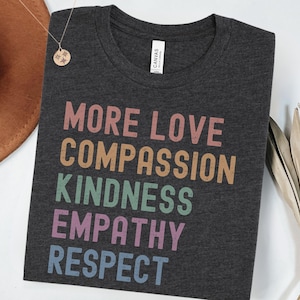 May include: A dark gray t-shirt with the words "MORE LOVE", "COMPASSION", "KINDNESS", "EMPATHY", and "RESPECT" printed in rainbow colors.