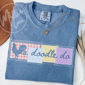 Könnte beinhalten: Ein blaues T-Shirt mit kurzen Ärmeln, das eine Grafik eines Hahns und den Text a doodle do zeigt. Das Design besteht aus vier quadratischen Feldern mit rot-karierten, blau-karierten, gelb-karierten und lila gepunkteten Mustern auf blauem Stoff.