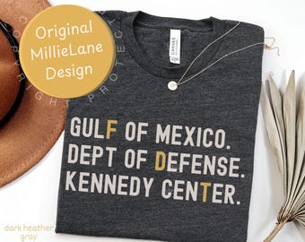 Könnte beinhalten: Ein dunkelgrau meliertes T-Shirt mit Rundhalsausschnitt und dem weißen und goldenen Text: GULF OF MEXICO. DEPT OF DEFENSE. KENNEDY CENTER. Das T-Shirt hat ein weißes Markenetikett am inneren Halsausschnitt und eine feine goldene Halskette.