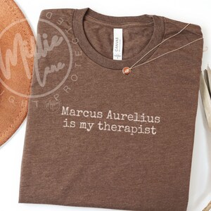 以下が含まれることがあります： 「Marcus Aurelius is my therapist」の白文字が入った茶色のTシャツ。丸いペンダントが付いたゴールドカラーのネックレスがシャツの上にあります。茶色の帽子が左上に写っています。