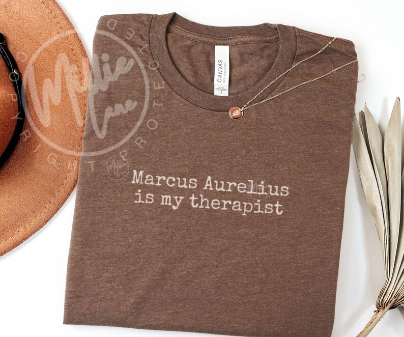 以下が含まれることがあります： 「Marcus Aurelius is my therapist」の白文字が入った茶色のTシャツ。丸いペンダントが付いたゴールドカラーのネックレスがシャツの上にあります。茶色の帽子が左上に写っています。