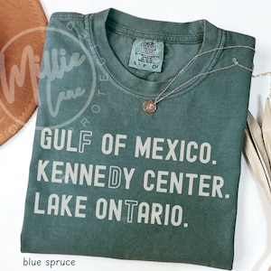May include: A blue spruce colored short-sleeve t-shirt with a crew neckline. The shirt features the printed text GULF OF MEXICO. KENNEDY CENTER. LAKE ONTARIO. in a light beige sans-serif font. A silver-toned necklace rests on the shirt collar.
