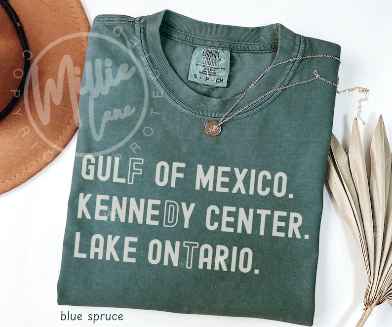 May include: A blue spruce colored short-sleeve t-shirt with a crew neckline. The shirt features the printed text GULF OF MEXICO. KENNEDY CENTER. LAKE ONTARIO. in a light beige sans-serif font. A silver-toned necklace rests on the shirt collar.