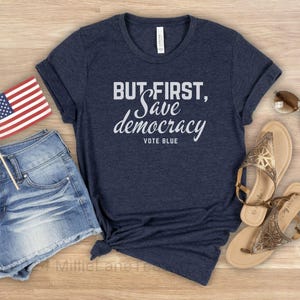 May include: A navy blue t-shirt with the slogan "BUT FIRST, Save democracy, VOTE BLUE". The image also shows a small American flag, denim shorts, and sandals.