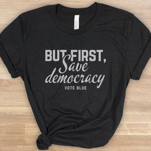 May include: Dark grey t-shirt featuring the text "BUT FIRST, Save democracy VOTE BLUE" in white and light grey. The sleeves are folded, and the bottom is knotted.