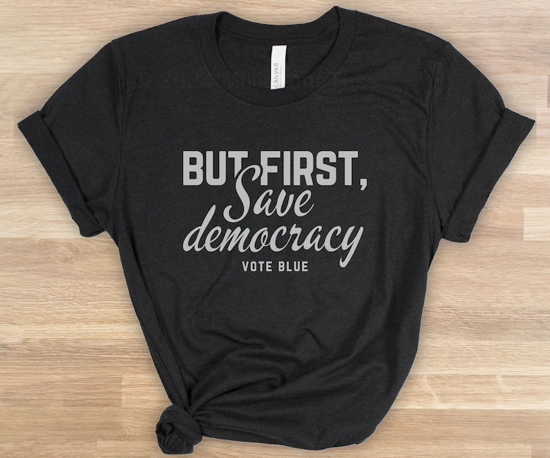 May include: Dark grey t-shirt featuring the text "BUT FIRST, Save democracy VOTE BLUE" in white and light grey. The sleeves are folded, and the bottom is knotted.