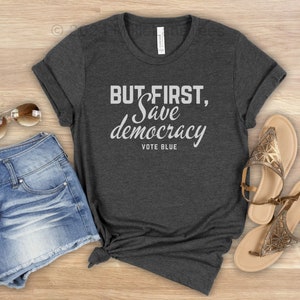 May include: A charcoal grey t-shirt featuring the white text "BUT FIRST, Save democracy VOTE BLUE". The image includes denim shorts, sunglasses, and gold sandals, suggesting a relaxed, everyday outfit.