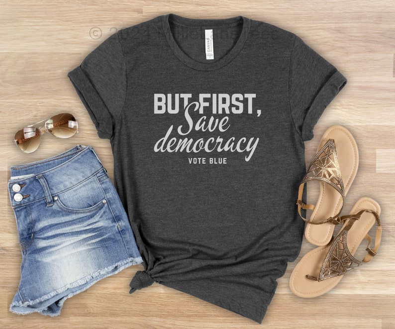 May include: A charcoal grey t-shirt featuring the white text "BUT FIRST, Save democracy VOTE BLUE". The image includes denim shorts, sunglasses, and gold sandals, suggesting a relaxed, everyday outfit.