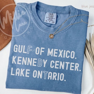 May include: A blue jean colored t-shirt with white text that reads GULF OF MEXICO. KENNEDY CENTER. LAKE ONTARIO. The shirt features a crew neckline and a Comfort Colors brand label on the inside collar. A gold-toned necklace rests on the fabric.