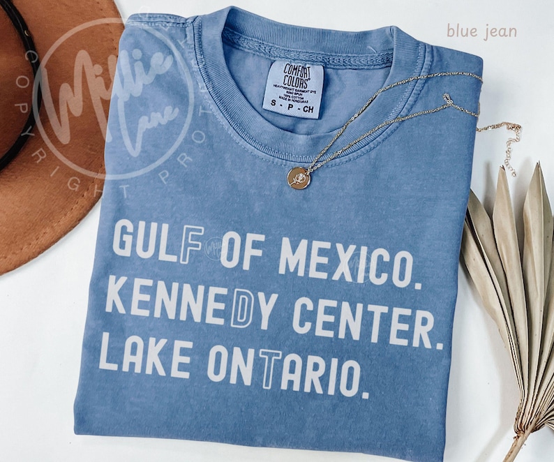 May include: A blue jean colored t-shirt with white text that reads GULF OF MEXICO. KENNEDY CENTER. LAKE ONTARIO. The shirt features a crew neckline and a Comfort Colors brand label on the inside collar. A gold-toned necklace rests on the fabric.
