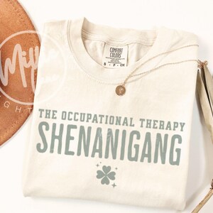 May include: Cream-colored t-shirt with the text "THE OCCUPATIONAL THERAPY SHENANIGANG" in gray-green letters. A four-leaf clover graphic is below the text. A gold necklace and a tan hat are also in the image.