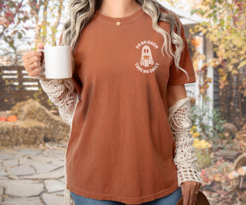 Peut inclure: Un t-shirt couleur rouille avec le texte "DO NO HARM TAKE NO SHEET" et un motif de fant&ocirc;me. Le t-shirt est &agrave; col rond et &agrave; manches courtes. La personne tient une tasse blanche et porte une manche en dentelle cr&egrave;me.