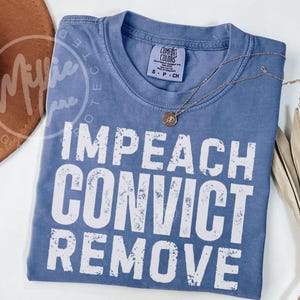 Könnte beinhalten: Ein blaues kurzärmeliges T-Shirt mit Rundhalsausschnitt und einem Etikett der Marke Comfort Colors. Das Kleidungsstück zeigt den weißen, im Used-Look gehaltenen Schriftzug IMPEACH CONVICT REMOVE in Großbuchstaben auf der Vorderseite.