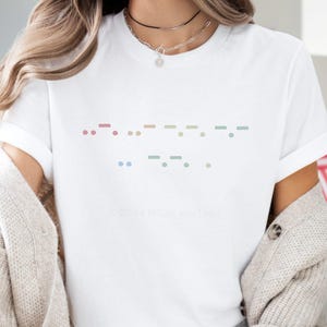 May include: White t-shirt with a Morse code design in pastel colors. The design features dots and dashes arranged in a horizontal pattern. The text "&copy; 2024 MilltownTees" is printed at the bottom of the shirt.