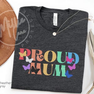 May include: Dark heather gray t-shirt with the words "PROUD MUM" in colorful letters and butterfly graphics. A gold necklace with a pendant is on the shirt. The shirt is on a white surface.