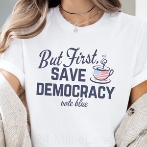 May include: White t-shirt with the text "But First, Save Democracy vote blue" in navy blue lettering. A coffee cup with an American flag design is above the text.